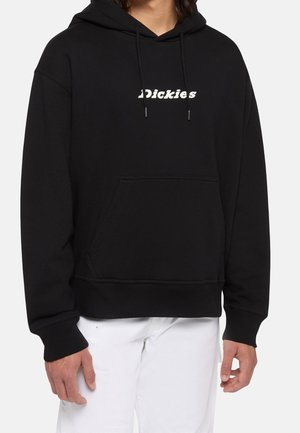 Czarna bluza z kapturem z białym logo "Dickies" na piersi, noszona z białymi spodniami, widok z przodu, ręce po bokach.