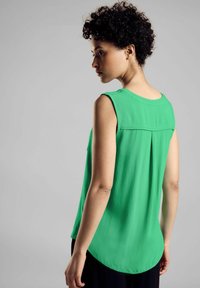 Vihreä hihaton paita, jossa on väljä malli, pyöreä pääntie ja takapuolella laskostus. Silkkinen kangas, kaareva helma, joka on yhdistetty tummiin housuihin.