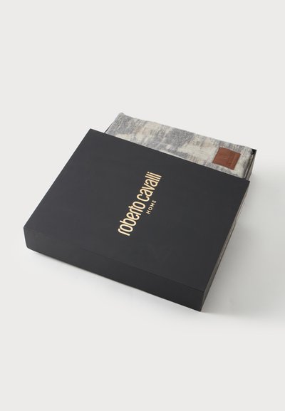 Μαύρο κουτί με χρυσό λογότυπο "roberto cavalli HOME" μερικώς ανοιχτό, αποκαλύπτοντας ένα διπλωμένο γκρι υφασμάτινο σχέδιο μέσα με δερμάτινη ετικέτα επωνυμίας.