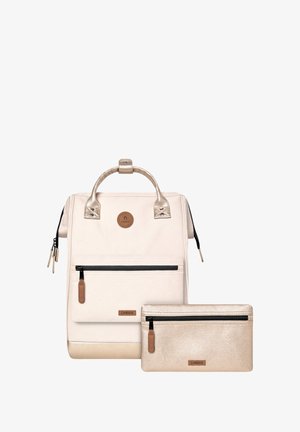 Sac à dos couleur crème avec des accents dorés brillants, comportant une poche zippée à l'avant et accompagné d'une petite pochette zippée dorée. Finition texturée.