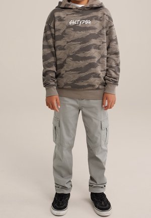 Barn iført grå og sort camouflagemønstret hættetrøje med teksten "SALTYDOG", lysegrå cargobukser og sorte sneakers, står med afslappede hænder.