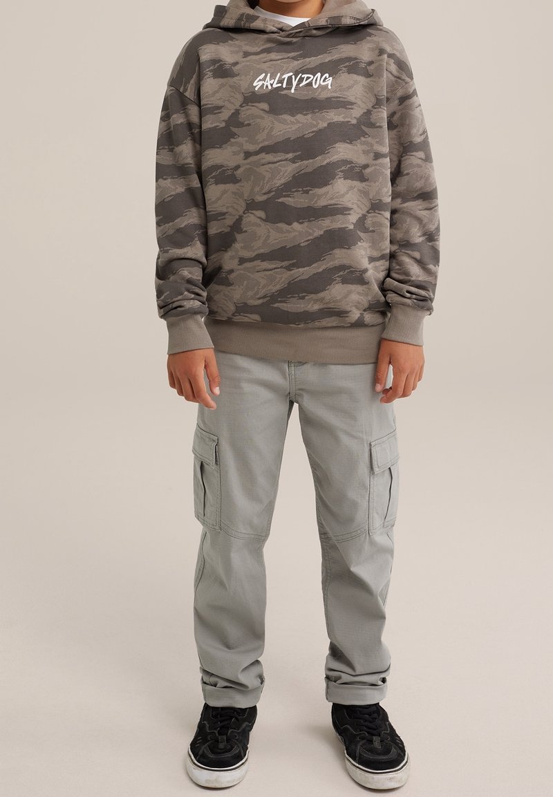 Dziecko ubrane w szarą i czarną bluzę z kapturem w kamuflażu z napisem "SALTYDOG", jasnoszare spodnie cargo i czarne tenisówki, stojące ze swobodnie opuszczonymi rękami.