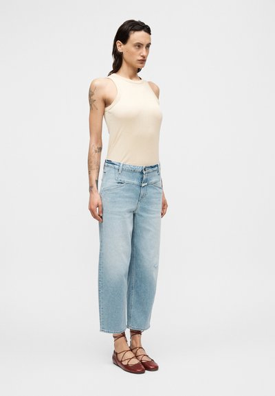 Haut sans manches beige, jean bleu clair à jambes larges, et chaussures plates bordeaux à lacets. Lignes épurées, style décontracté, avec coutures visibles et légère usure.