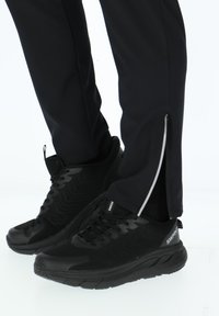 Person trägt schwarze Sportschuhe und schwarze Hosen mit seitlichen Reißverschlüssen und steht vor einem weißen Hintergrund.