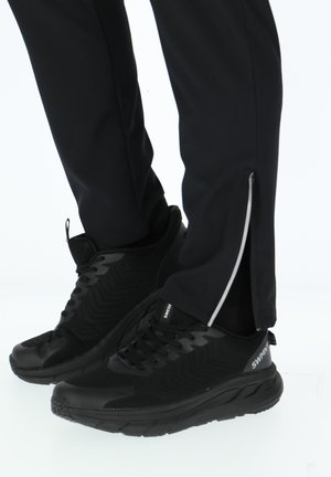 Person iført sorte sportssko og sorte bukser med sidelommer med lynlås, stående mod en hvid baggrund.