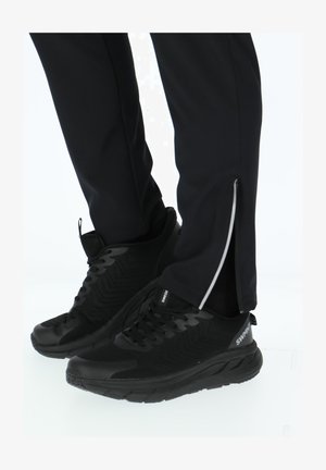 Person iført sorte sportssko og sorte bukser med sidelommer med lynlås, stående mod en hvid baggrund.