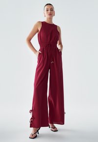 Bordozna kombinezonka brez rokavov, z elastičnim pasom, nastavljivim vrvico, širokimi hlačnicami in stranskimi žepi. Poudarjeno z vezalkami na robu.