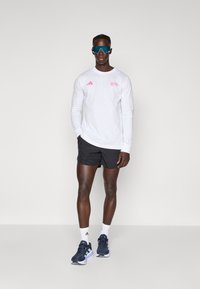 White long-sleeve shirt with pink logo, black shorts, navy sneakers, and blue sunglasses. Smooth fabric and athletic fit.