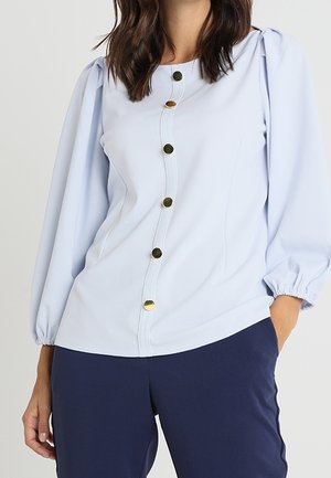 Kobieta w jasnoniebieskiej bluzce z długim rękawem zapinanej na złote guziki z przodu oraz w granatowych spodniach, z ręką w kieszeni, na jednolitym tle.