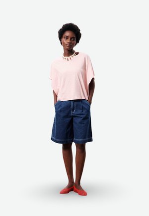 Femme debout, les mains dans les poches, portant une chemise ample rose clair, un short en jean bleu à hauteur des genoux, des chaussures plates rouges et un collier de coquillages.