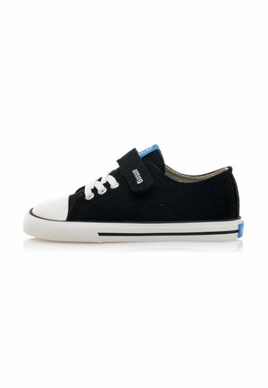 Zapatilla deportiva negra de corte bajo con puntera de goma blanca, cordones blancos, tira de velcro y suela blanca con franja negra.