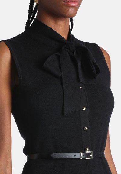 Top noir sans manches avec un col noué en forme de nœud, des boutons dorés sur le devant et une ceinture noire fine à la taille.