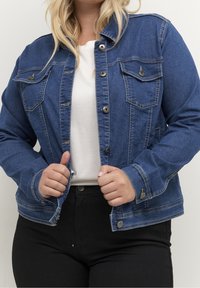 Denimjakke i mørkeblå med knaplukning, spids krave, brystlommer og kontrastsyning. Båret over en hvid top.