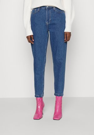 Personne portant un jean taille haute bleu et des bottines roses vives, debout devant un fond blanc uni.