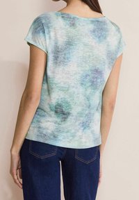 Kvinna i kortärmad, ljusblå mönstrad topp och mörkblå jeans sedd bakifrån mot en beige bakgrund.