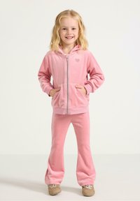 Růžový vlněný teplákový komplet s propínací mikinou, která má detail srdce, doplněný o rozšířené kalhoty. Měkká textura a neformální styl.