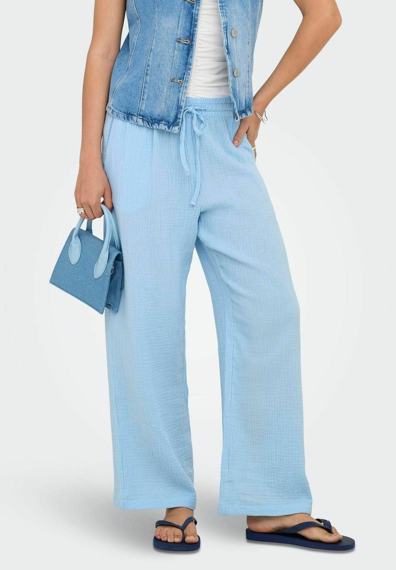 Svijetloplave široke hlače od teksturiranog materijala, uparene s traper prslukom, držeći malu plavu torbicu; nosi tamnoplave havajke.