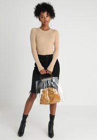Sac en toile transparent avec une pochette à cordon brune à l'intérieur, porté par un mannequin en haut à côtes beige et jupe noire, sur un fond neutre.