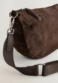 Braune Wildledertasche mit runder Form, Reißverschlussverschluss und metallischen Beschlägen. Wird mit einem strukturierten braunen Gurt geliefert, der mit einem silbernen Clip befestigt ist.