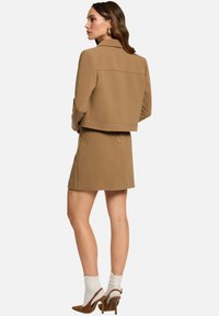 Chaqueta ajustada marrón con falda a juego, ambas de tejido texturizado. Presenta un cuello y detalles de cremallera, combinada con zapatos de tacón y calcetines.