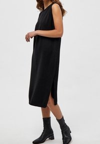 Abito midi nero senza maniche in morbido tessuto a maglia, con fessure laterali e un design semplice e svasato. Abbinato a stivaletti neri alla caviglia.