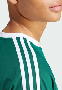 Green jersey with white horizontal stripes on the shoulders, featuring a crew neck and a soft, textured fabric. Close-up view of the neckline.