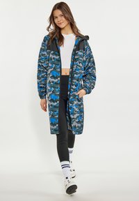 Jachetă lungă cu model camouflage în nuanțe de albastru, gri și negru. Are fermoar în față, manșete elastice și glugă. Purtați-o peste un top alb scurt și colanți.