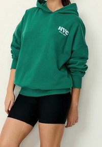 Persona vestida con una sudadera verde con el texto "NYC" y pantalones cortos ajustados negros, de pie contra una pared clara y lisa.