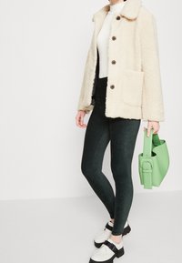 Donna che indossa una giacca sherpa crema, un dolcevita bianco, leggings verde scuro e mocassini bianchi con suole nere, mentre tiene una borsa leggera verde.