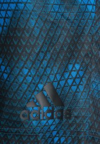 Adidas atletisk tyg med ett svart och blått geometriskt mönster, med en framträdande svart logotyp på en texturerad bakgrund.