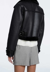 Veste courte en simili cuir noir avec des détails en boucle portée sur un pull côtelé blanc et un short à carreaux par une personne debout.