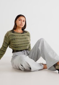Crochet oliivivihreä lyhythihainen neule, jossa on avoin kudontamalli, yhdistettynä vaaleanharmaisiin, löysiin verkkareihin ja yksinkertaisiin jalkineisiin.