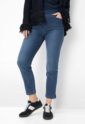 Person iført blå cropped jeans, sorte og hvide sneakers og en sort tekstureret cardigan med frynser på ærmerne, stående mod en hvid baggrund.
