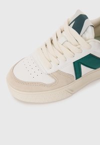 Sneaker bianca e beige con accenti verdi, caratterizzata da una punta in suede testurizzato, perforazioni per la traspirabilità e lacci spessi.