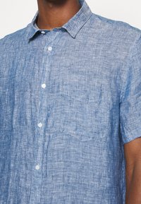 Camicia azzurro chiaro a maniche corte in lino; presenta un colletto a punta, una tasca sul petto e un tessuto a trama con un motivo sottile.