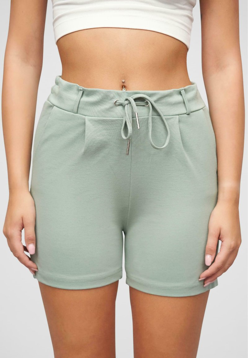 Lysegrønne, elastiske shorts med snor i livet, sidelommer og rullede kanter. Stoffet virker blødt med en glat tekstur.