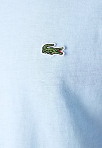 Polo de color azul claro con un tejido suave, que presenta un logo bordado de cocodrilo verde con acentos rojos en el lado izquierdo.