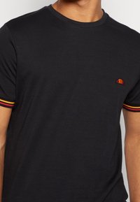 Black cotton t-shirt with short sleeves, featuring red and yellow striped accents on cuffs and a small embroidered logo.
