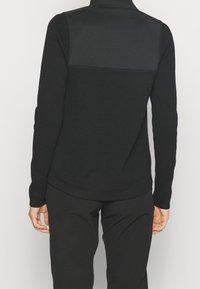 Haut noir à manches longues présentant un tissu texturé avec un panneau lisse dans le haut du dos, un col montant et une coupe ajustée.