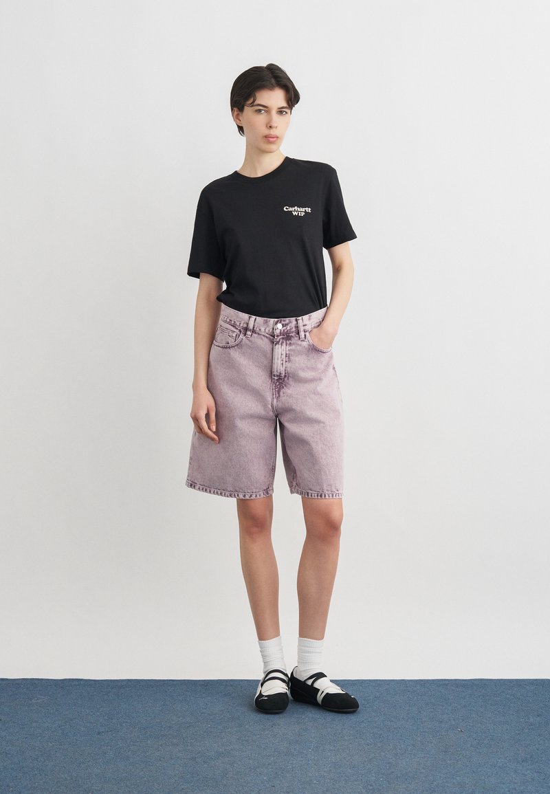 Junge Person steht und trägt ein schwarzes Carhartt WIP T-Shirt, helllila Jeansshorts, weiße Socken und schwarze Schuhe auf blauem Boden.