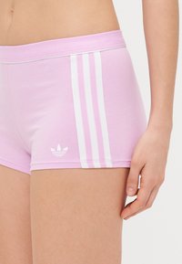 Gaiši rozā šorti, kas izgatavoti no mīksta auduma, ar baltu triju svītru dizainu sānos un nelielu baltu logotipu uz apakšmala.