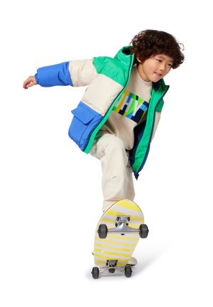 Enfant portant une doudoune verte, bleue et écrue, un pantalon beige et un pull avec un logo coloré, en équilibre sur un skateboard rayé jaune.