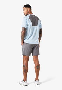 T-shirt de sport bleu clair avec un panneau arrière gris foncé, associé à un short gris. Présente des tatouages sur les jambes et des baskets blanches.