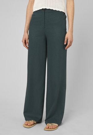 Femme portant un pantalon large vert foncé, un haut sans manches en maille crème et des sandales beiges avec un motif de feuilles.