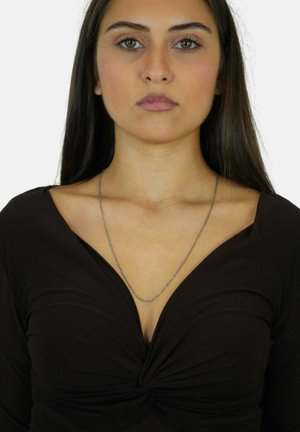 Donna con capelli lunghi e scuri che indossa un top nero a scollo a V e una sottile collana d'argento su uno sfondo bianco.