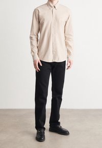 Beige overhemd met lange mouwen en een button-down kraag, voorzien van een geborduurd logo, gecombineerd met zwarte broek en zwarte loafers.