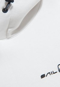 Tejido blanco con una textura suave que presenta un escote redondo y un logo negro de "SAIL" en la parte inferior derecha, junto con un detalle de cordón.