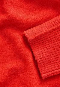 Tessuto di maglia rossa con una texture liscia e polsini a costine, caratterizzato da un aspetto morbido e fluffy e un colore uniforme in tutto il tessuto.