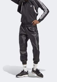 Schwarze Sportjacke mit Reißverschluss und weißen Streifen, kombiniert mit schwarzen Jogginghosen aus Kunstleder. Schwarze Sneakers und weiße Socken mit sichtbarem Logo.