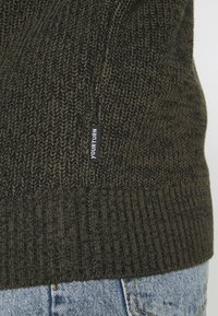 Tmavě zelený pletený svetr s žebrovaným lemem, zdobený strukturovaným vzorem a malou černou štítkem s nápisem "YOUR TURN" na boku.